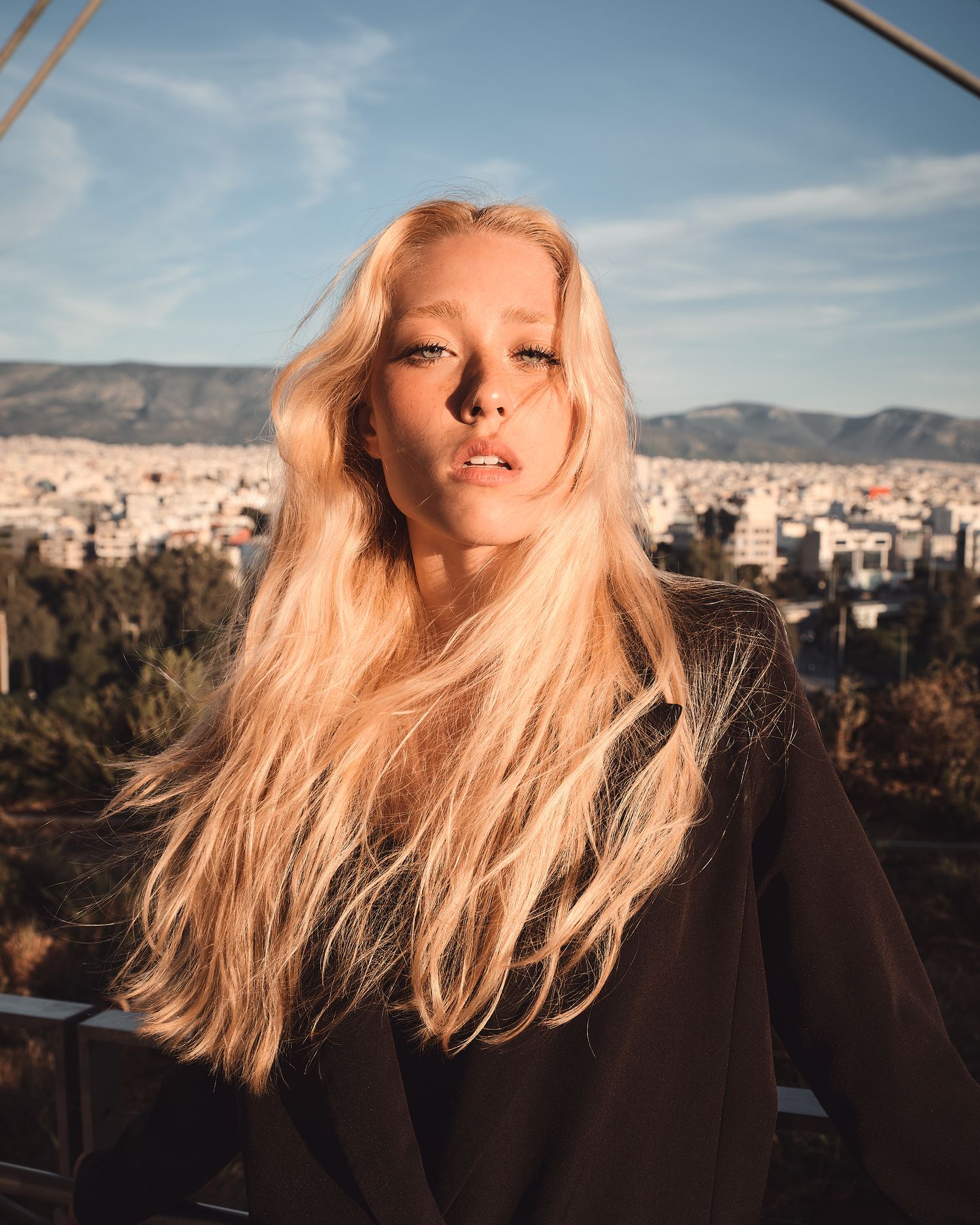 Blonde woman in black top poses against city skyline and mountains under blue sky.