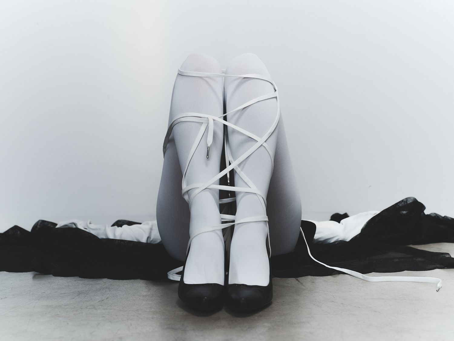 Person sitting with legs crossed wearing white lace-up ballet shoes on a light gray floor.