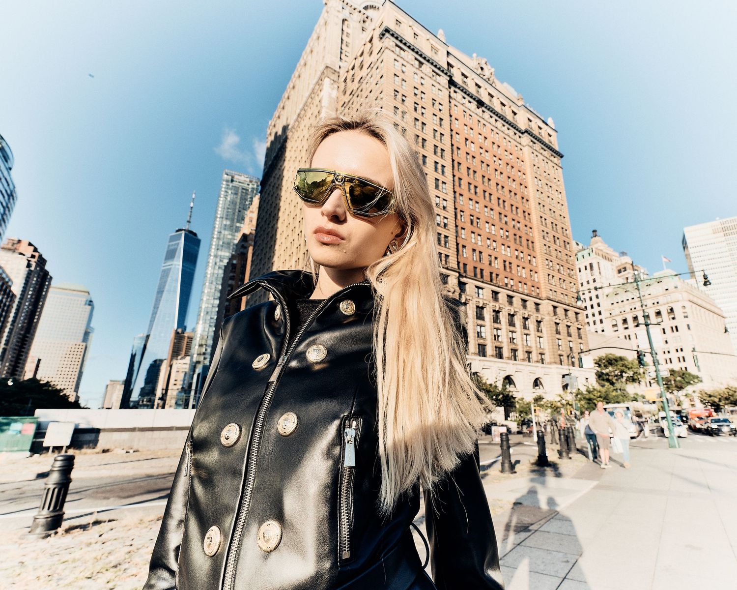 Blonde woman in black leather jacket and gold sunglasses poses on a sunny New York City street with skyscrapers behind her.