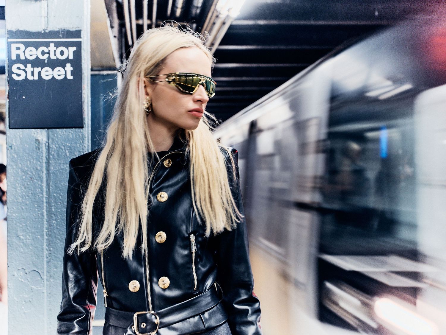 Blonde woman in black leather jacket with gold buttons poses at Rector Street NYC subway station.