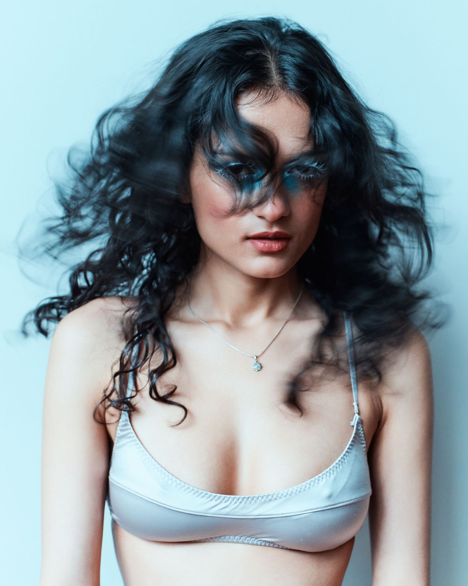 Young woman with long dark curly hair wearing a silver bralette and delicate necklace against a light blue background.