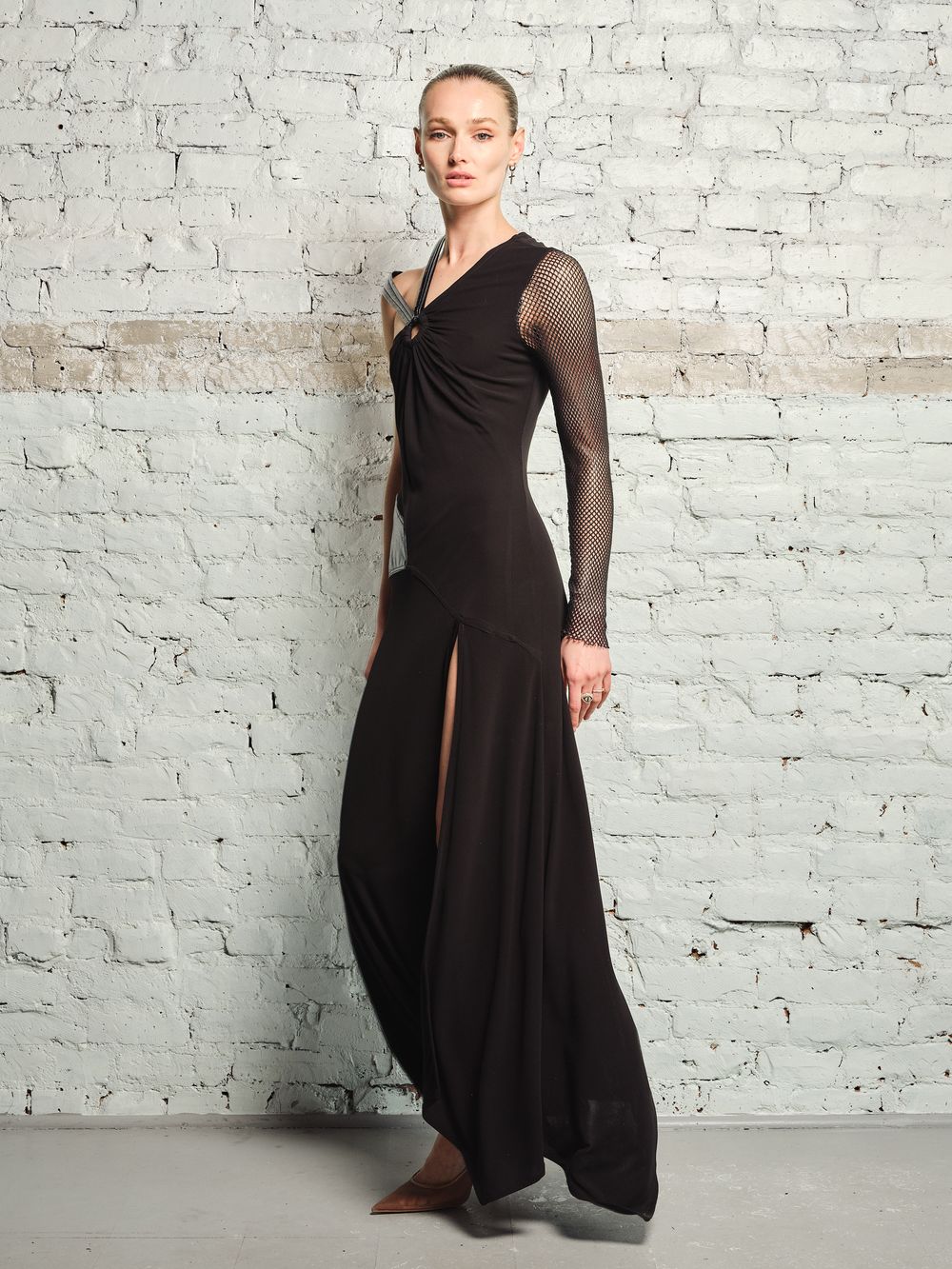 Model wearing a flowing black dress with asymmetrical hem and sheer long sleeves poses against textured brick wall.