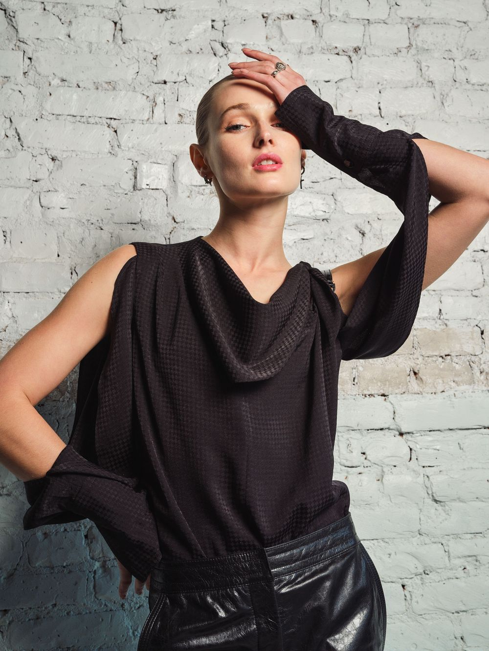 Fashion model in black cowl neck sweater and leather belt poses dramatically against white brick wall in series.
