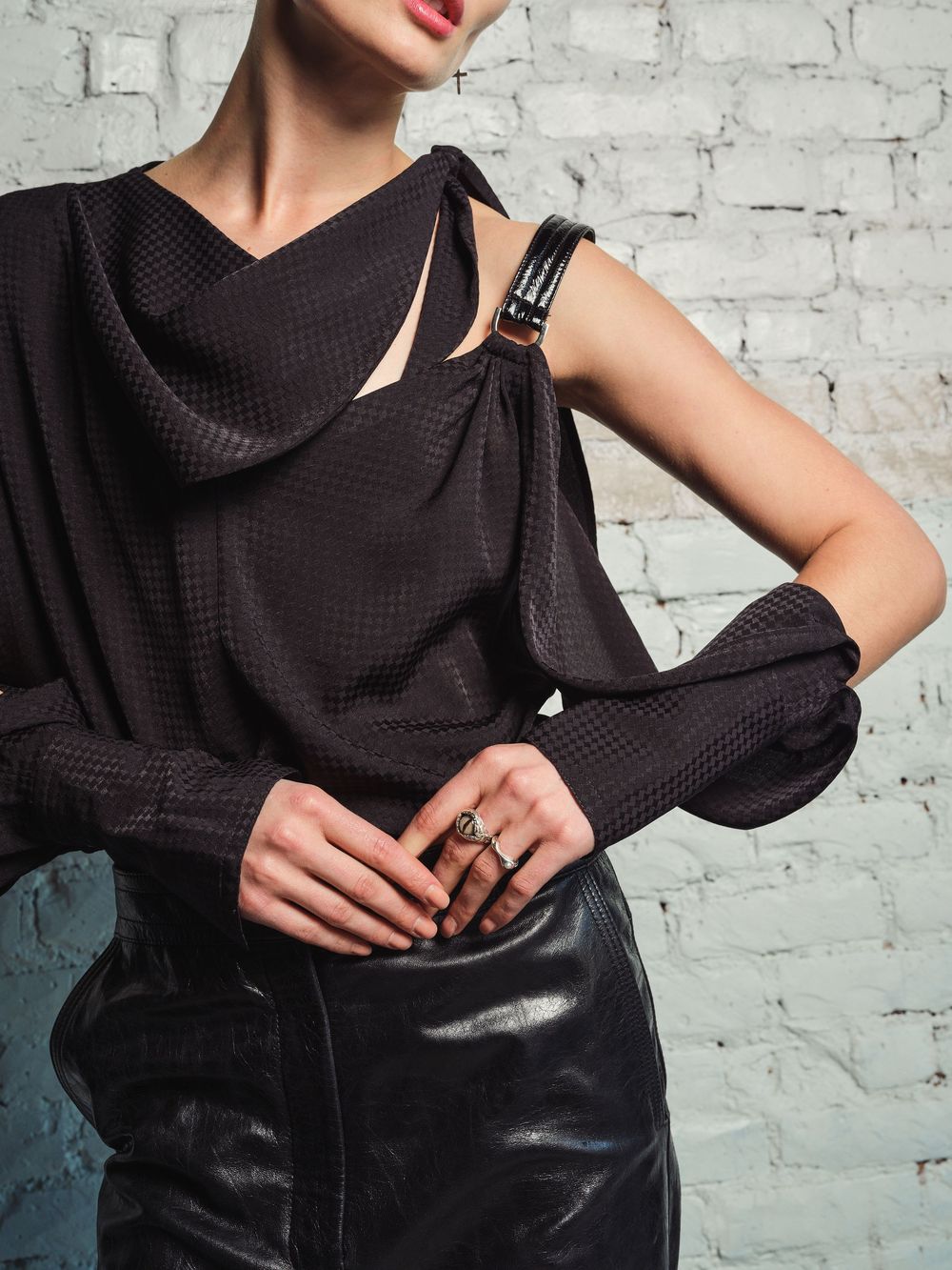 Closeup fashion details of black draped top with asymmetrical neckline against white brick background.