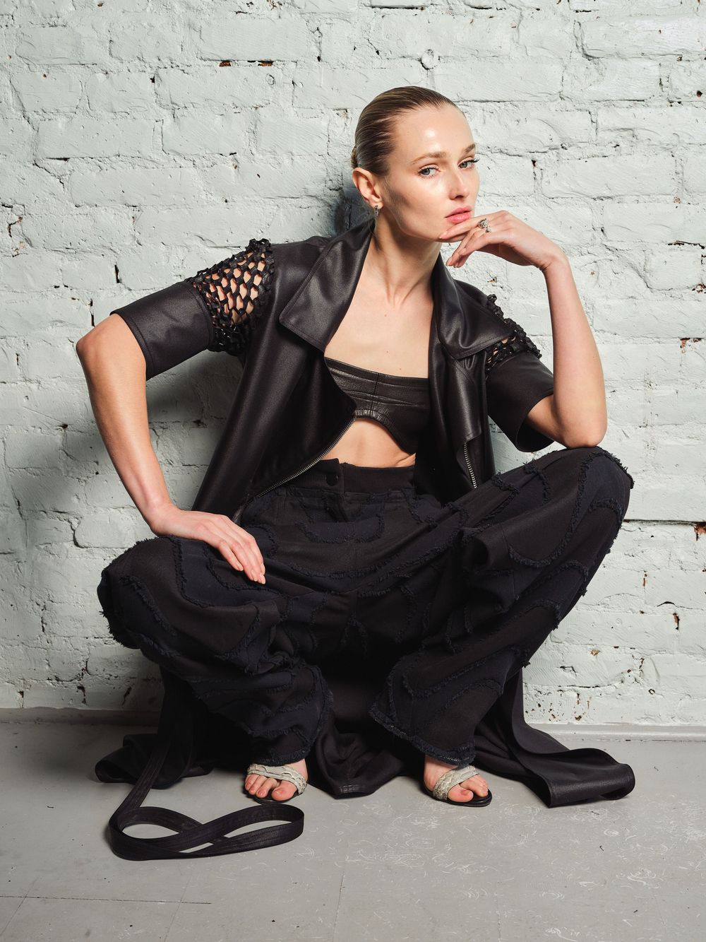 Model in black outfit reclining elegantly against white brick wall.
