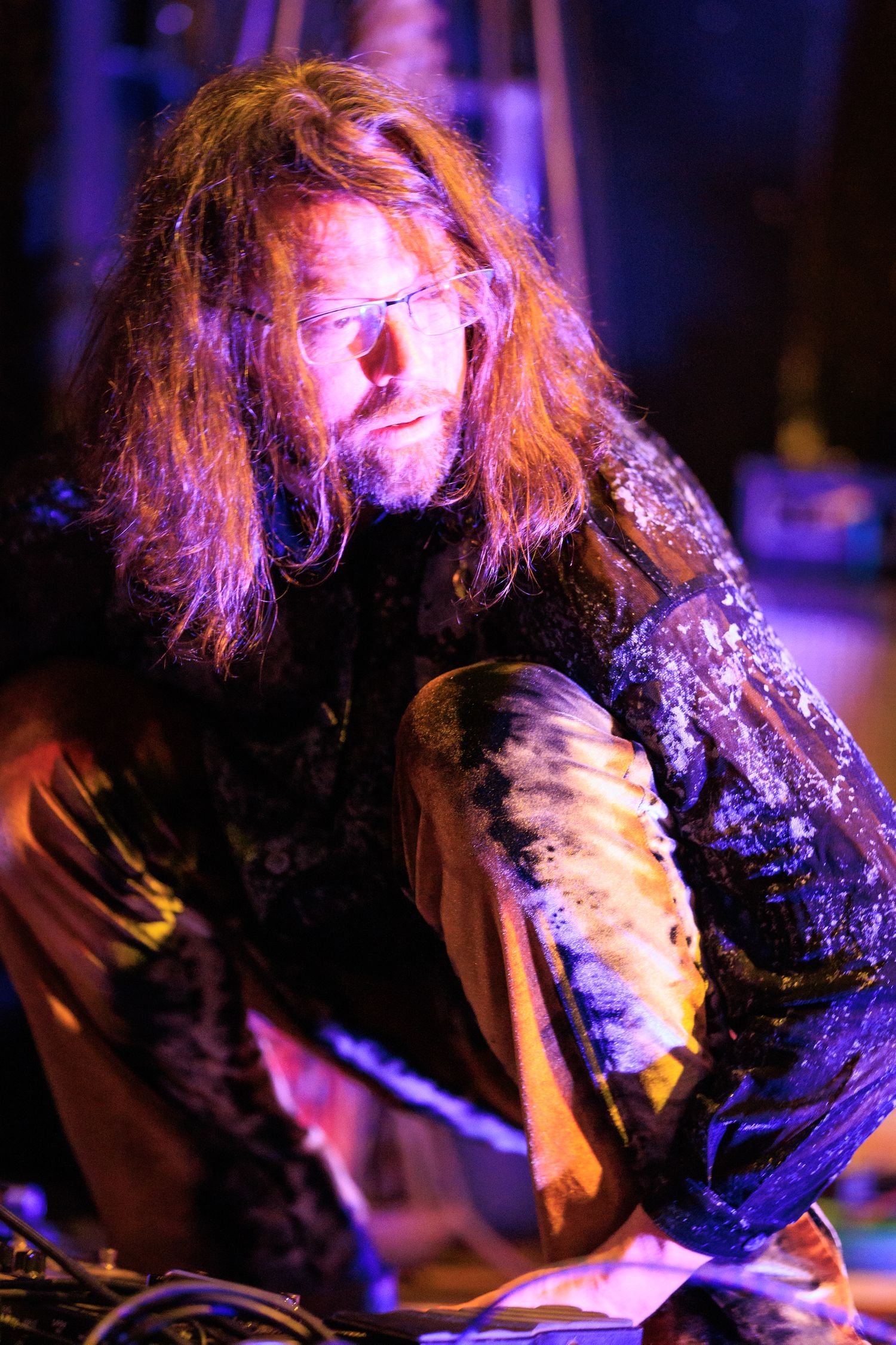 Long-haired bearded man with paint-covered clothing working under colorful purple stage lighting.