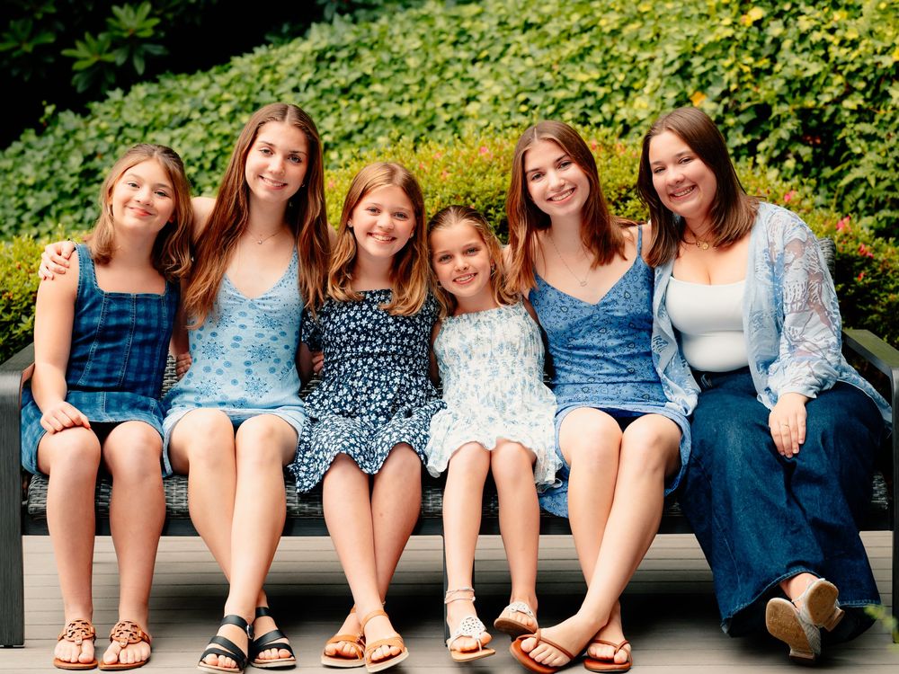 Row of people sitting casually on concrete ledge outdoors with green bushes in background.
