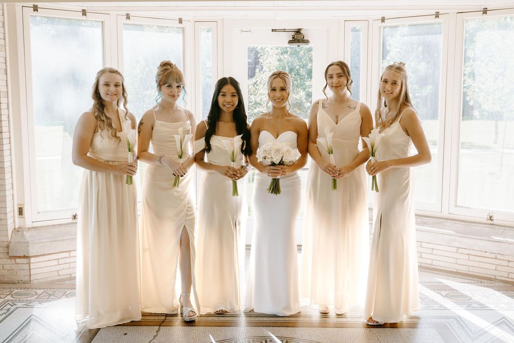 A bridal party in flowing white and cream dresses poses together in a sun-filled room.