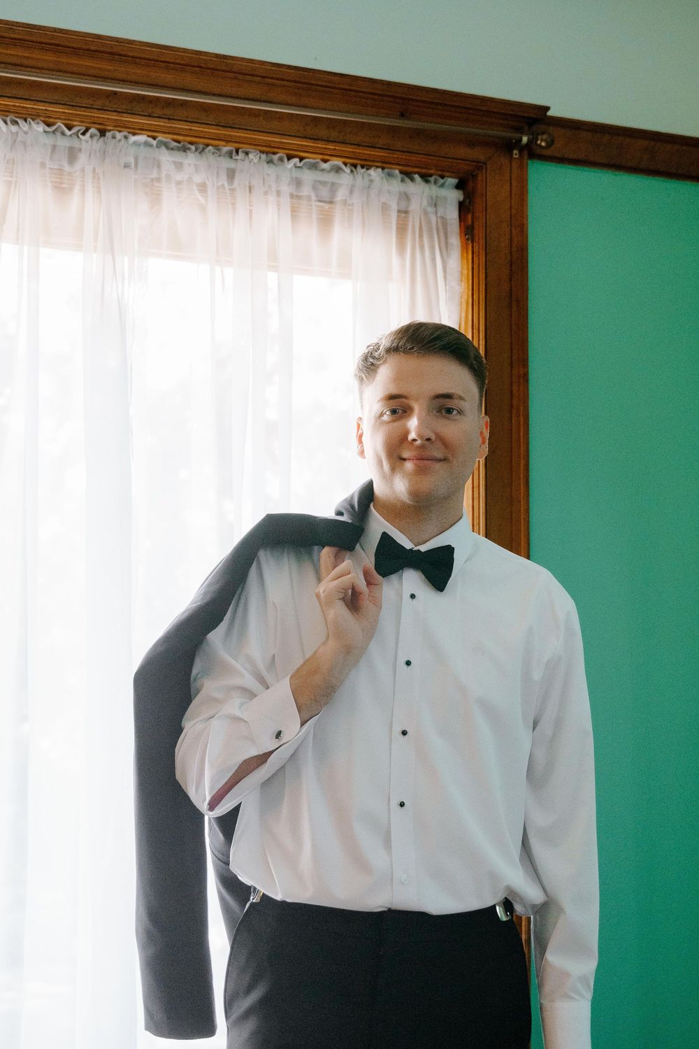 Person in formal white dress shirt and black bow tie poses in front of green wall and window with wooden trim.