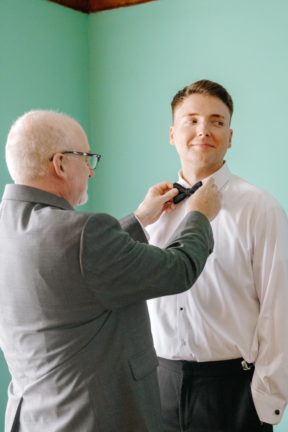 An older gentleman in grey helps adjust the bowtie of a younger person in formal attire.