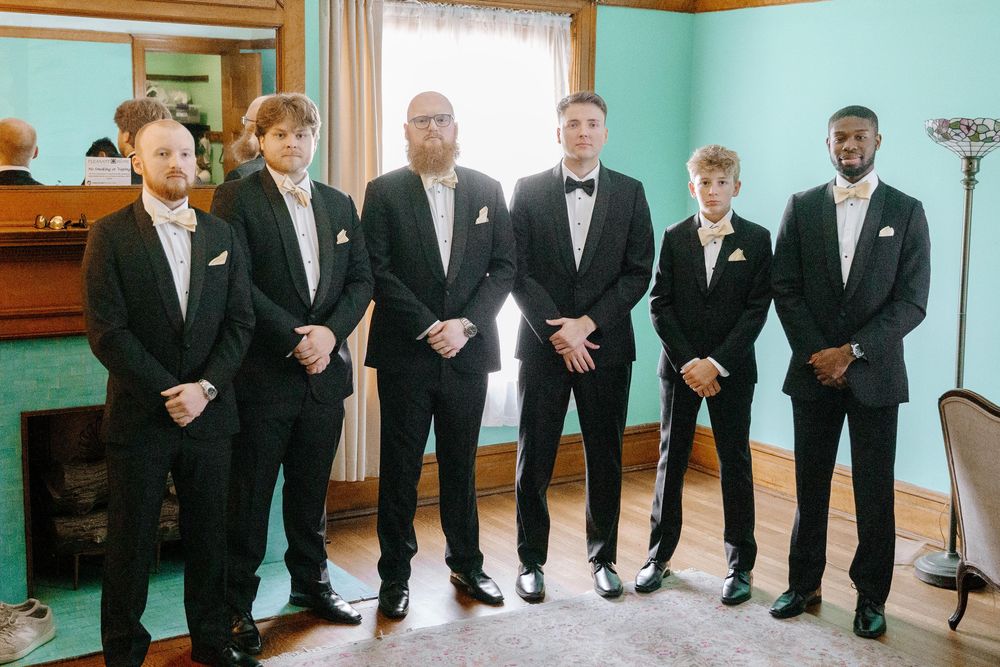 A line of groomsmen in classic black tuxedos stand together in a room with mint green walls.