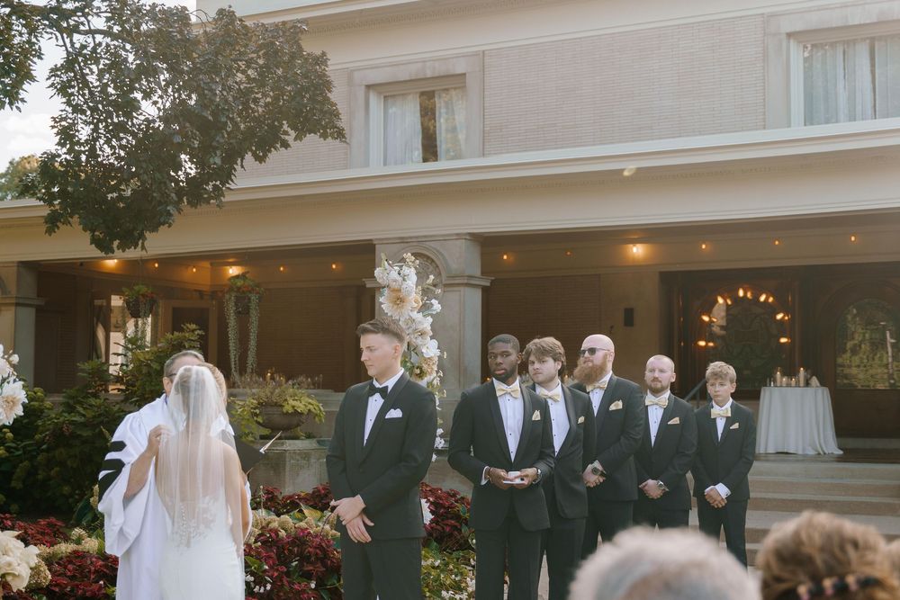 A wedding party stands in formation on the steps of an elegant white building during an outdoor ceremony.