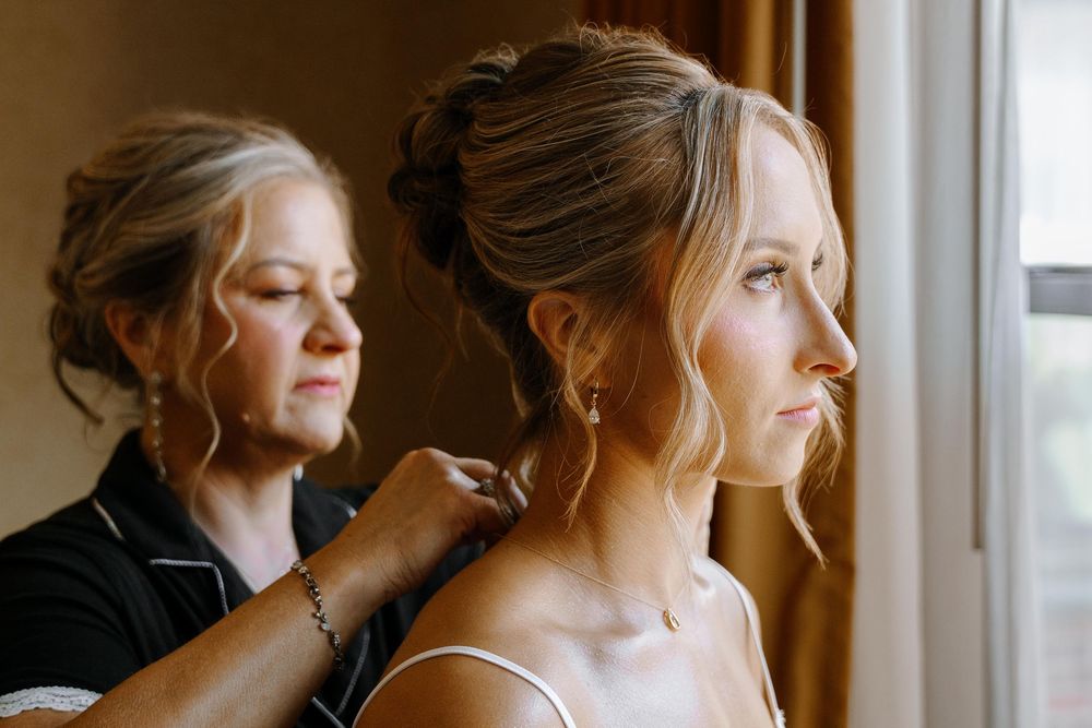 Bride in elegant dress gazes out window with soft natural light during wedding day preparation.