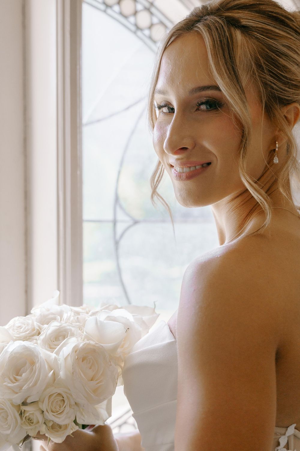Bride in strapless wedding dress smiles softly while holding white rose bouquet near window with natural light.