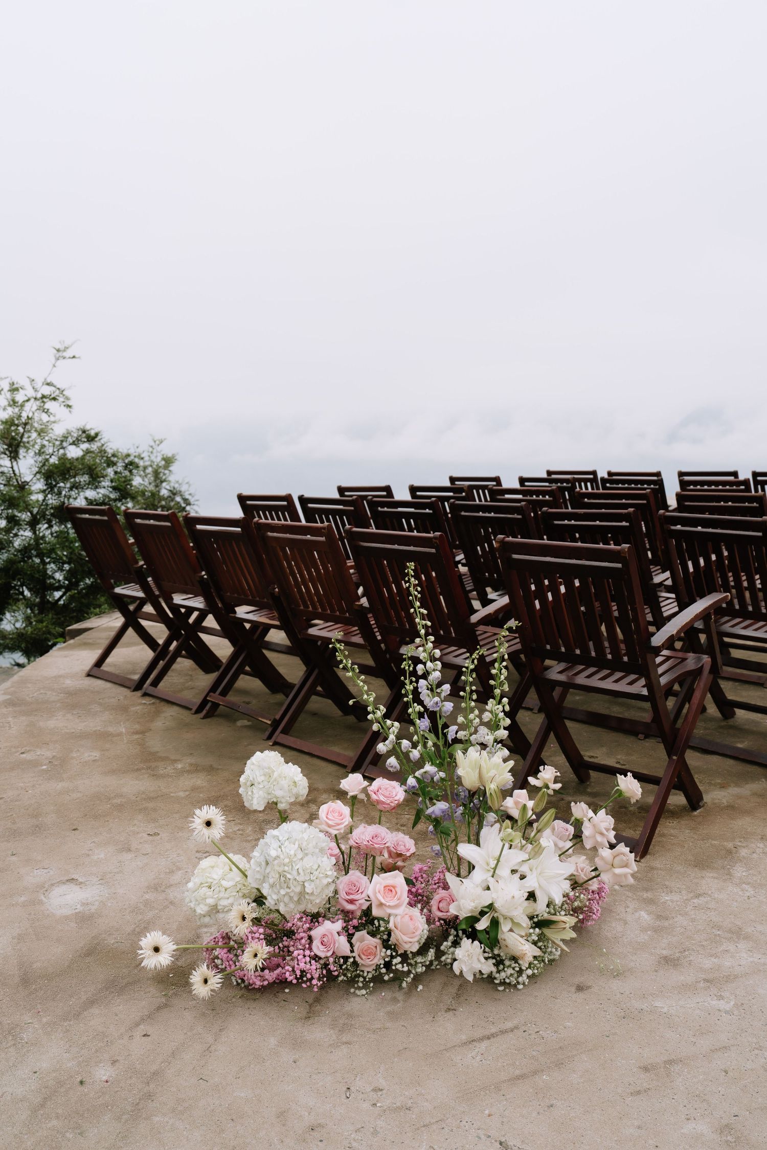 Pink and white floral arrangement beside rows of dark wooden chairs at an outdoor wedding ceremony.