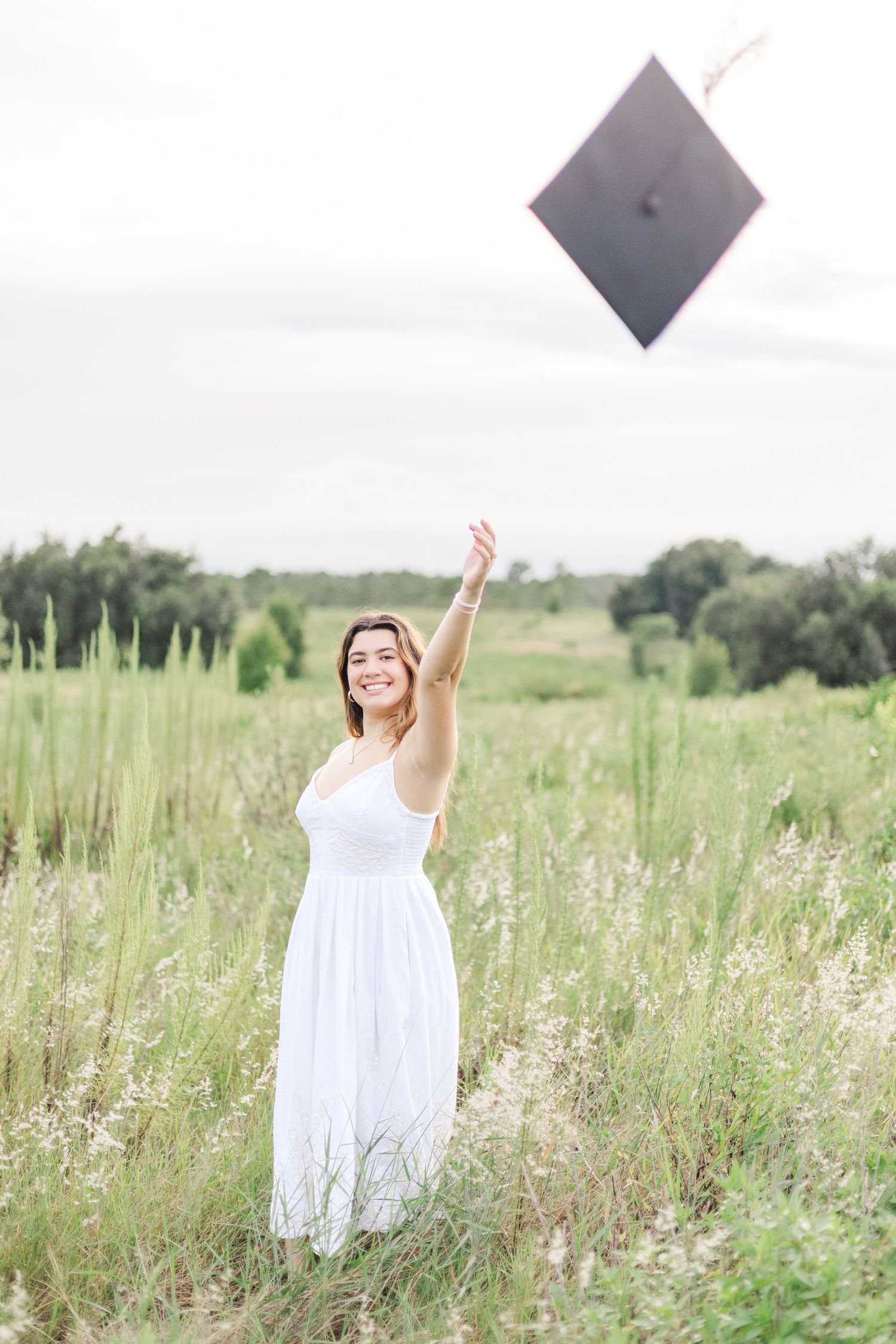 A group in white dresses poses playfully in a sunny meadow with tall grass and trees in the background.