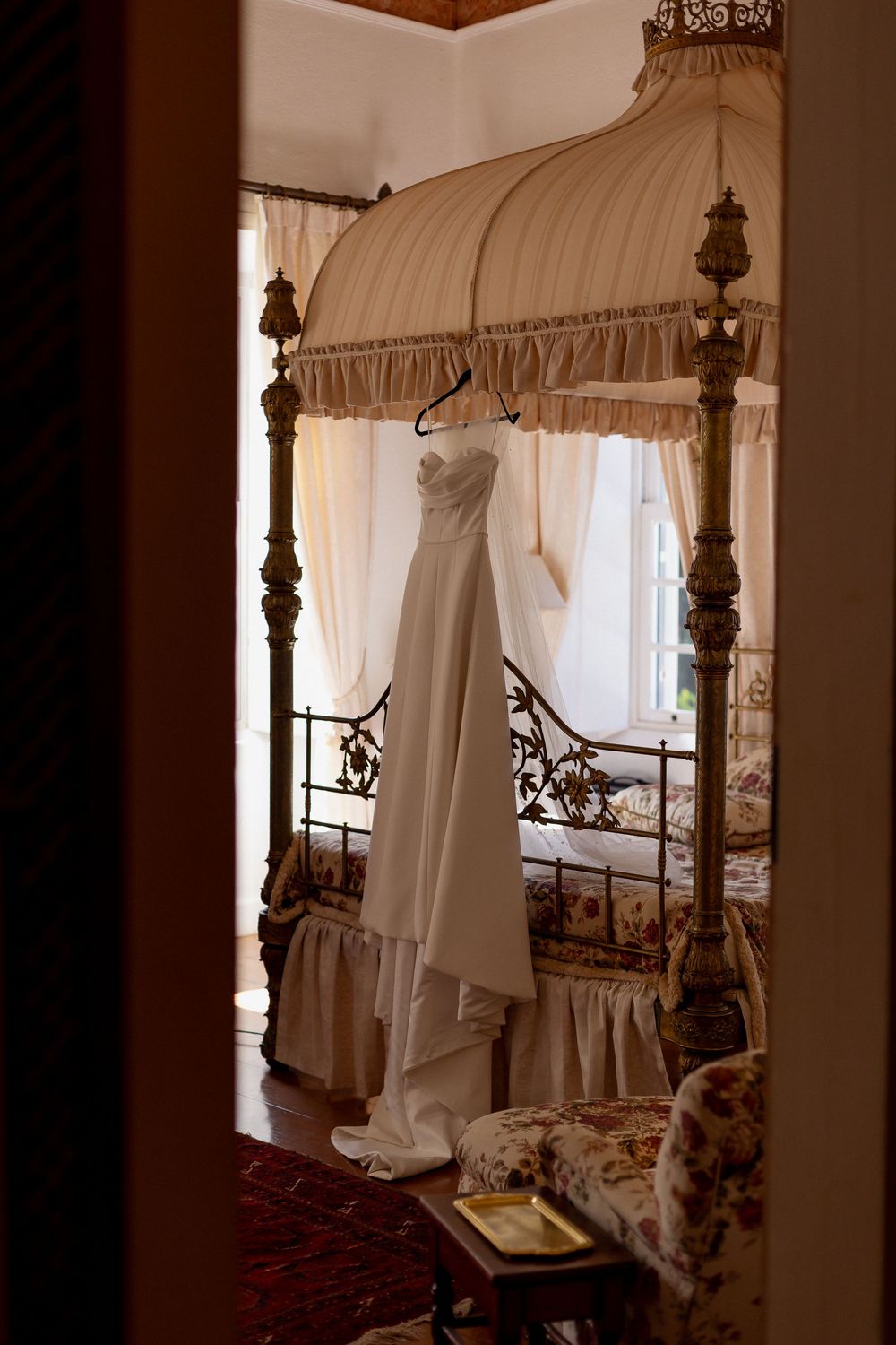An elegant white wedding dress hangs from an ornate canopy bed with antique wooden posts and luxurious draping.