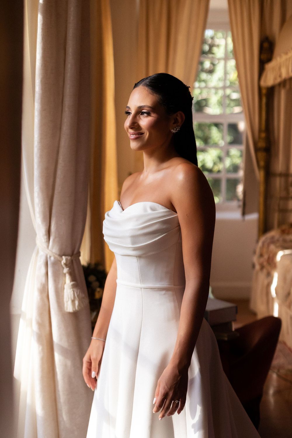 A bride in a strapless white wedding gown stands in soft window light creating an elegant silhouette in multiple poses.