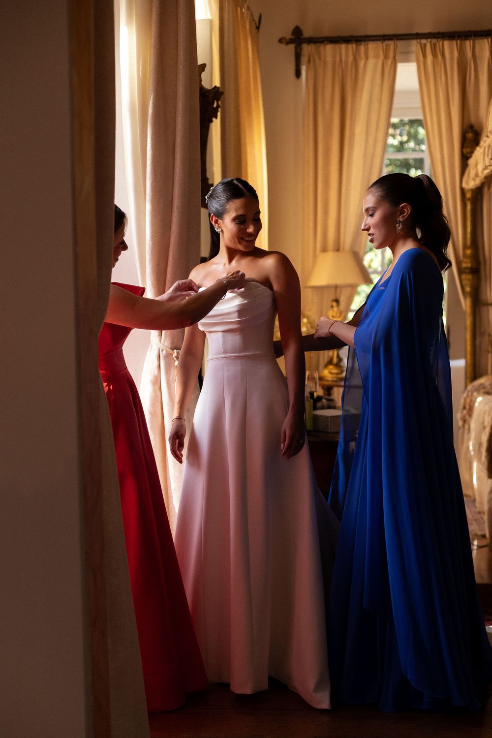 Three women in formal gowns - red, pink, and royal blue - gathered together in an elegant room with curtains.