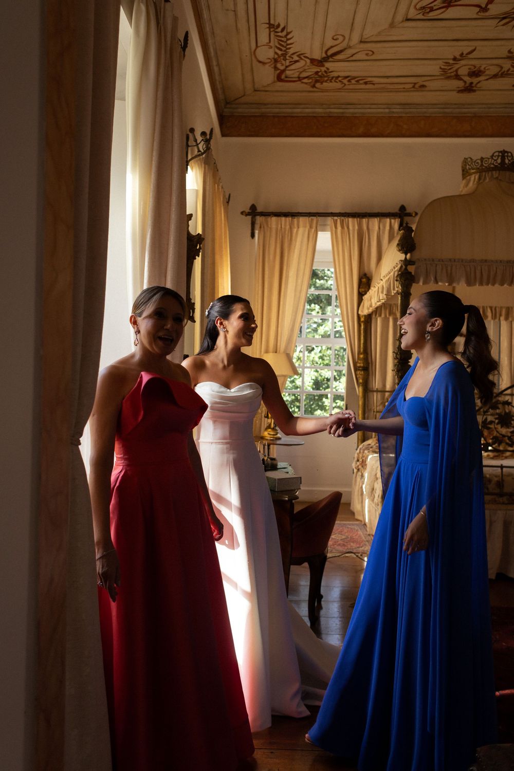 Three women in elegant evening gowns - red, white and blue - stand in conversation by a sunlit window in an ornate room.