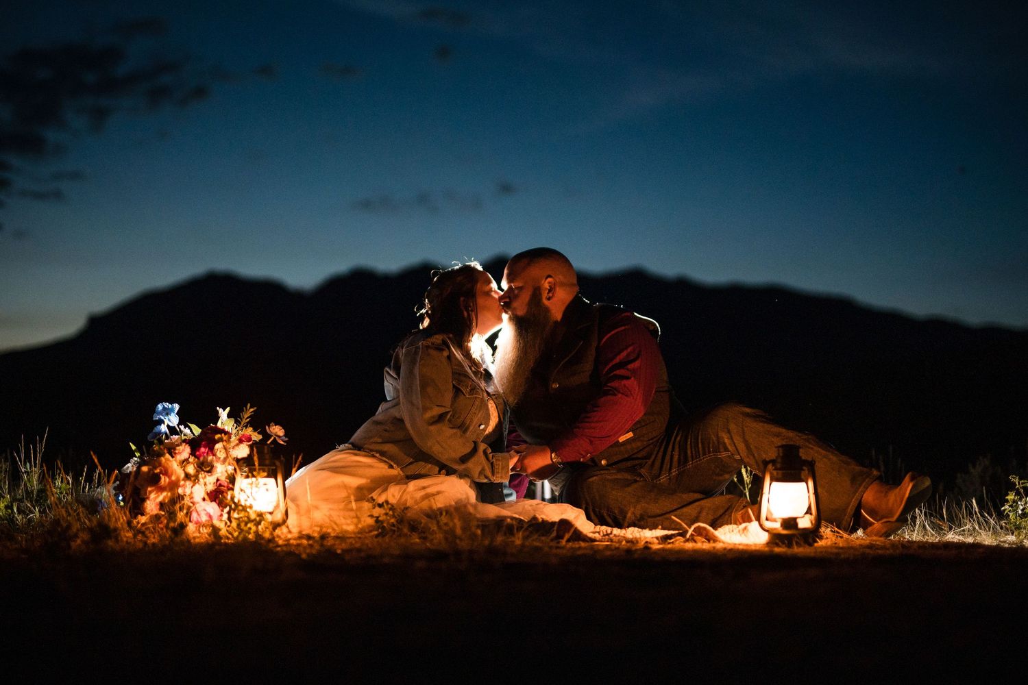 Glowing orange tent illuminates a dark mountain landscape with scattered camping gear and lights in the background.
