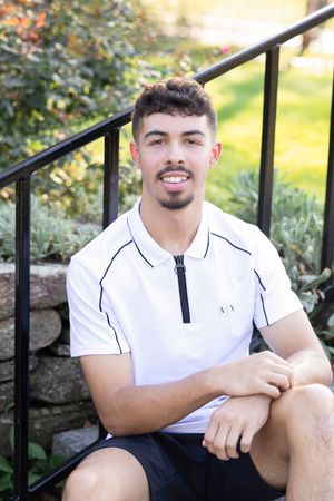 Young person in white polo shirt sitting outdoors on stone steps with greenery in background.