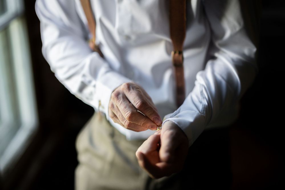 Close-up of someone adjusting brown leather suspenders over a crisp white dress shirt.