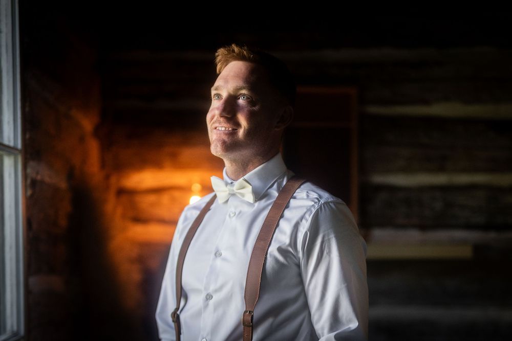 A groom in a white shirt and suspenders stands in dramatic window lighting.