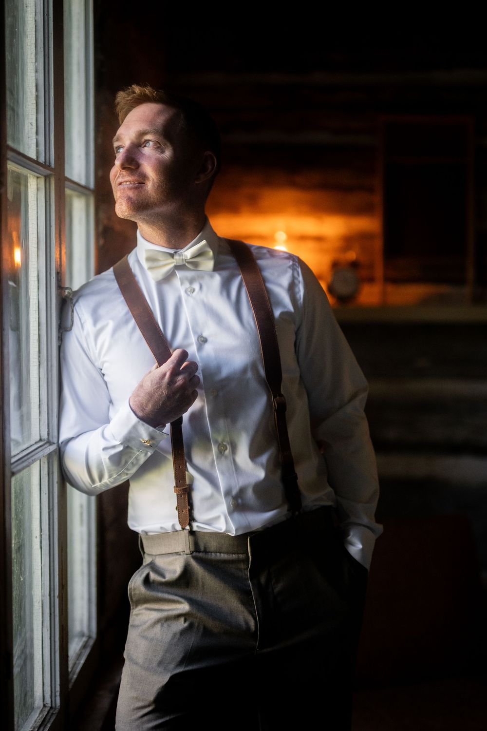 Groom in white shirt and suspenders poses by window with dramatic lighting.