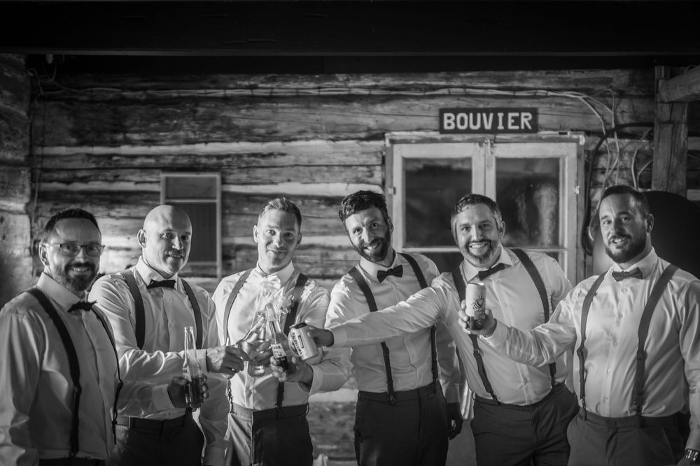 Groomsmen in suspenders and bowties pose together in front of rustic wooden walls.