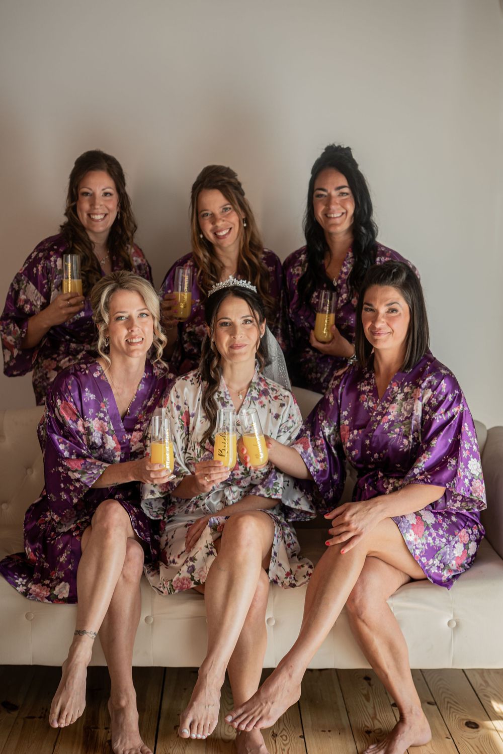 Group wearing matching purple floral robes pose while sitting on wooden floor during wedding preparations.