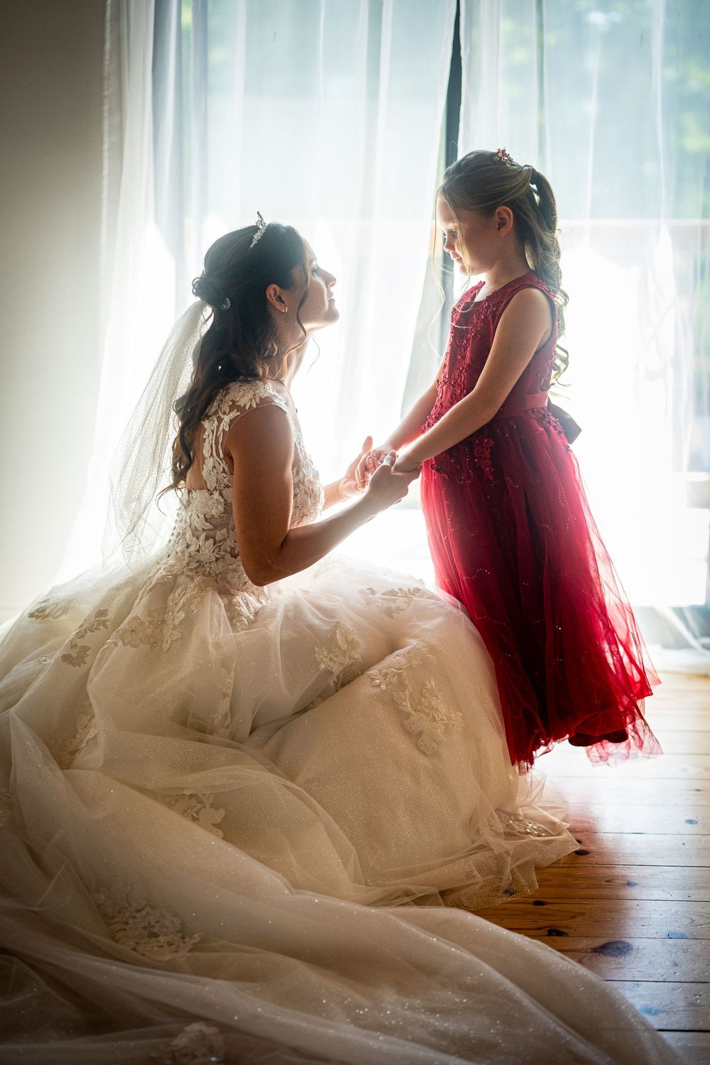 Tender moment between bride in lace gown and young attendant in red dress by window.