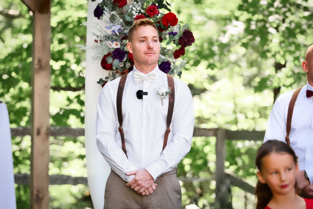 Wedding attendant in white shirt and suspenders stands under floral arch during outdoor ceremony.