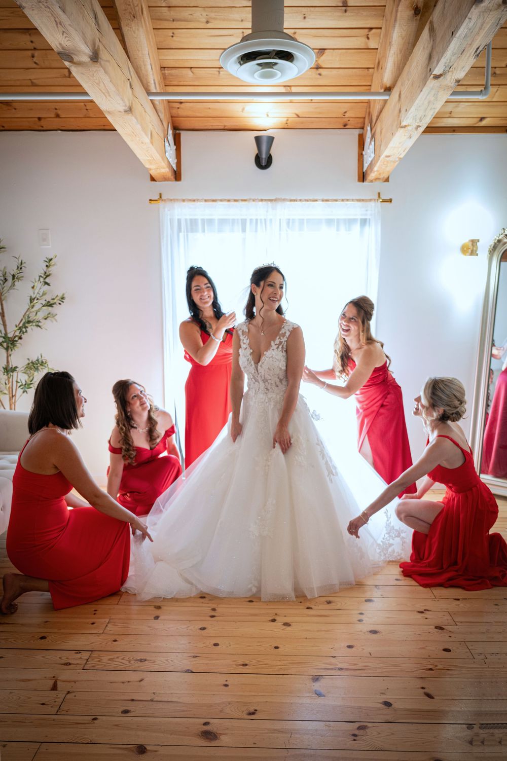Bridesmaids in red dresses pose playfully with a bride in a white gown on rustic wooden floors.