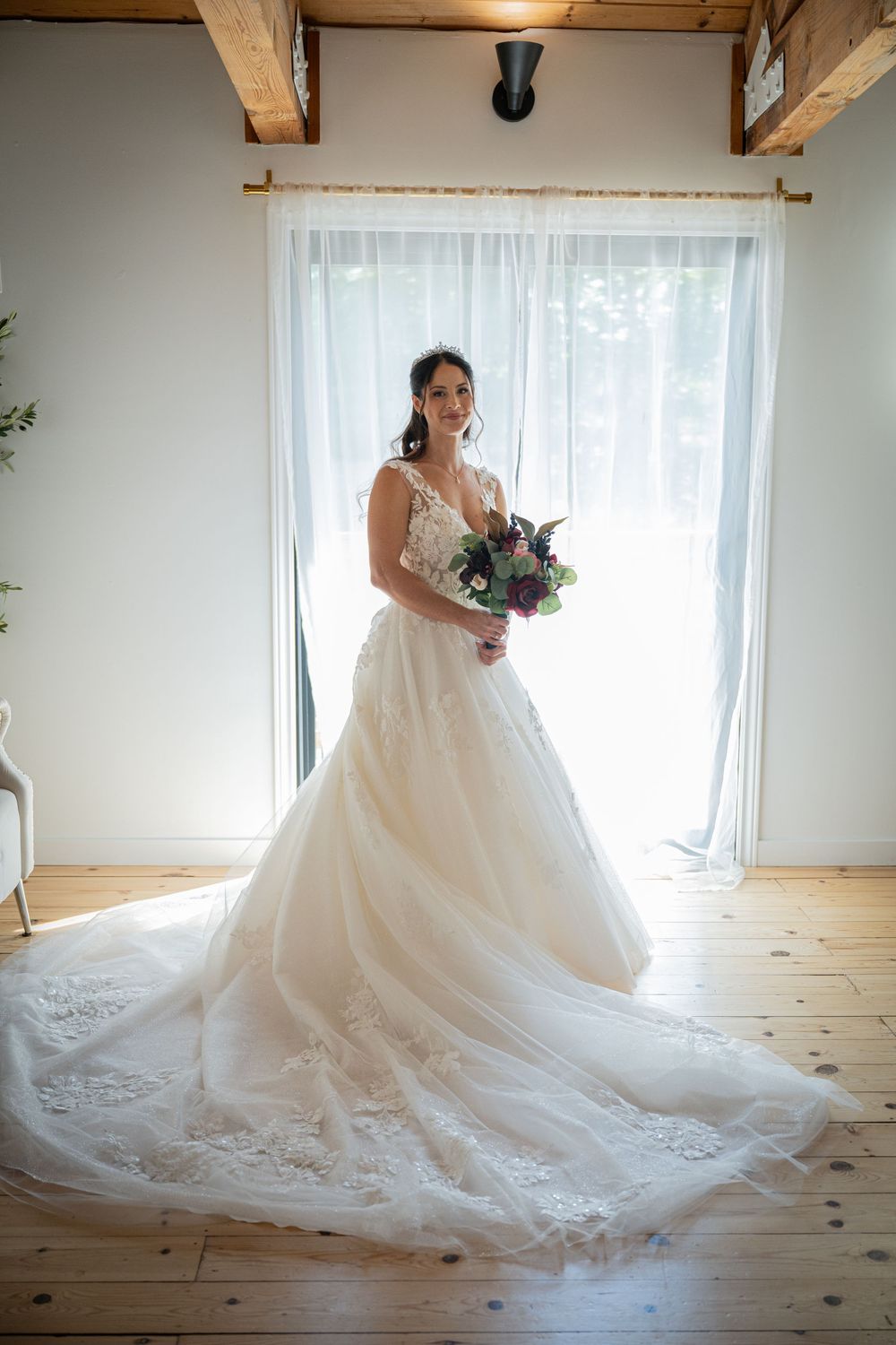 Elegant wedding dress with flowing tulle train captured in natural light against white windows and wooden flooring.