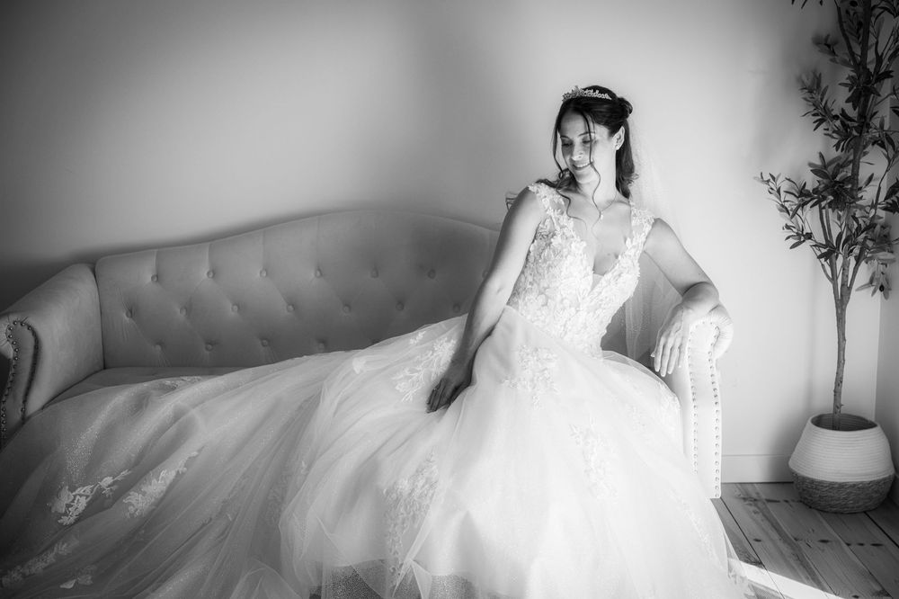 A bride in an elegant white ballgown sits gracefully on a tufted gray couch in a light-filled room.