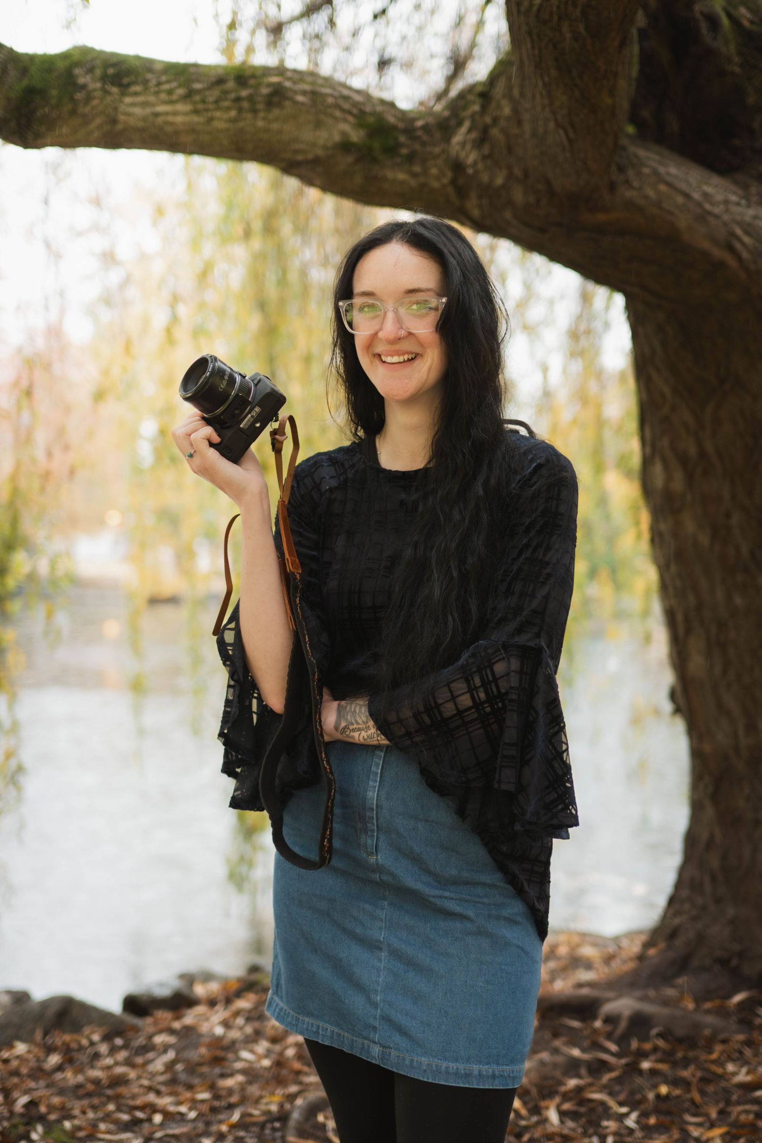 Smiling female photographer holding camera near autumn lake under large tree.