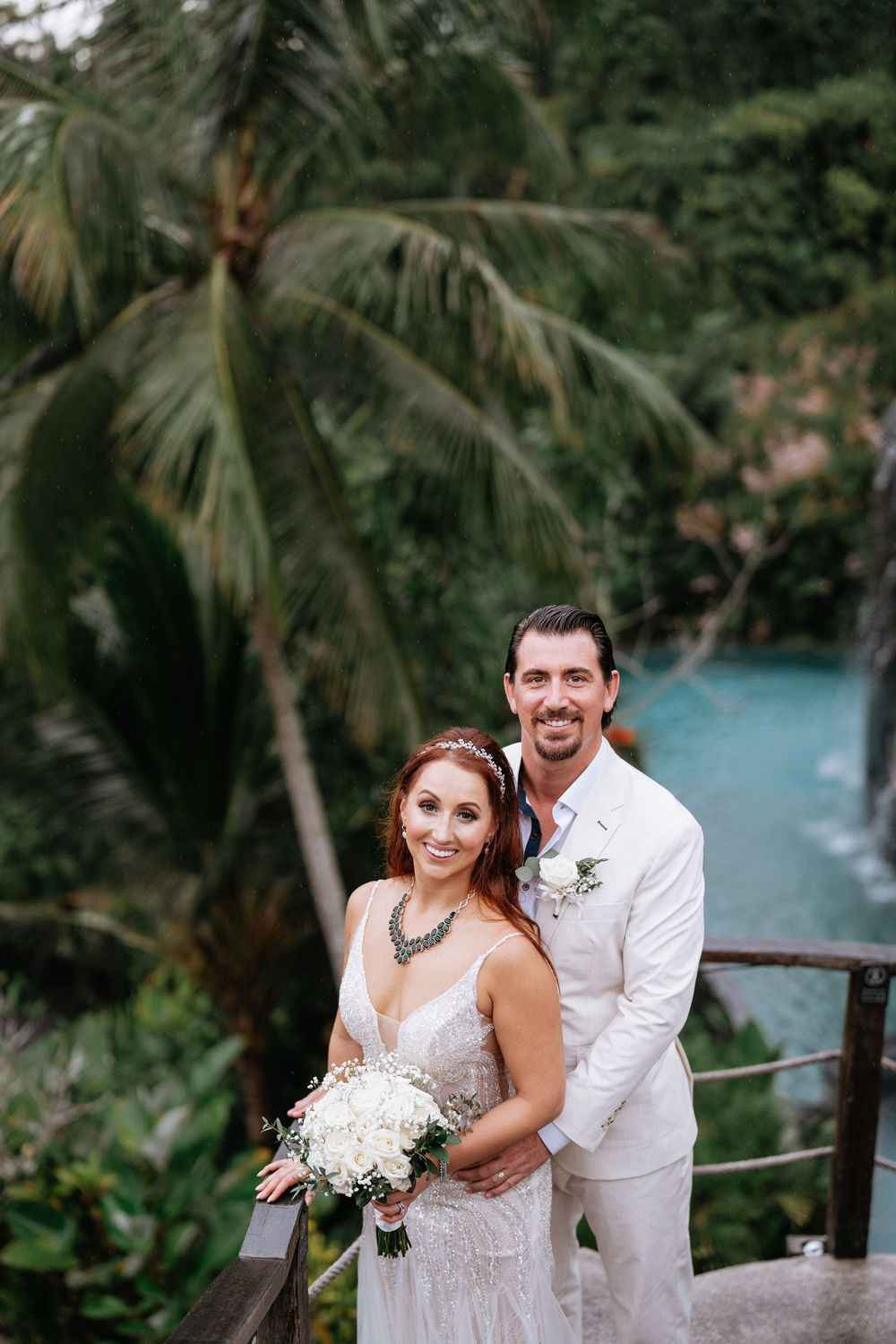 Wedding couple posing on stone steps with palm trees and turquoise water in background.