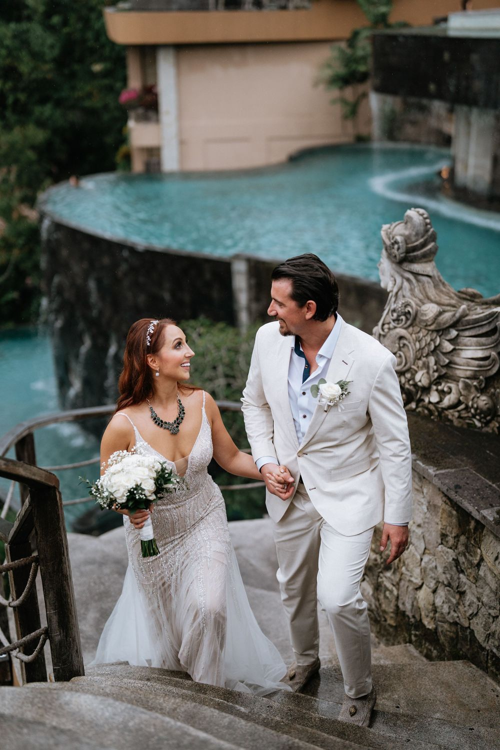 Wedding couple sharing romantic moments on stone steps overlooking tropical pool setting.