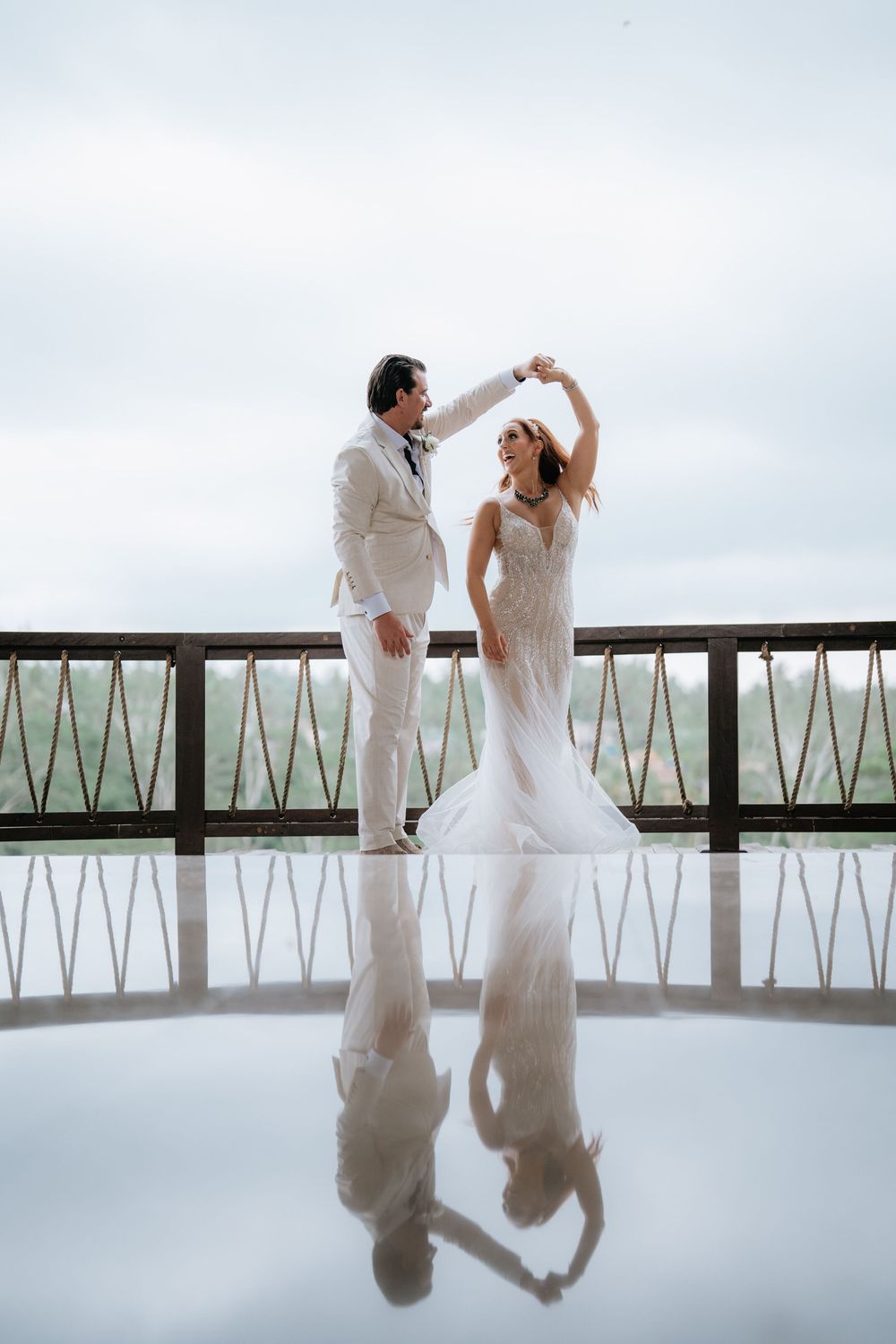 Couple in white attire dancing against minimalist railing with reflection on glossy floor.