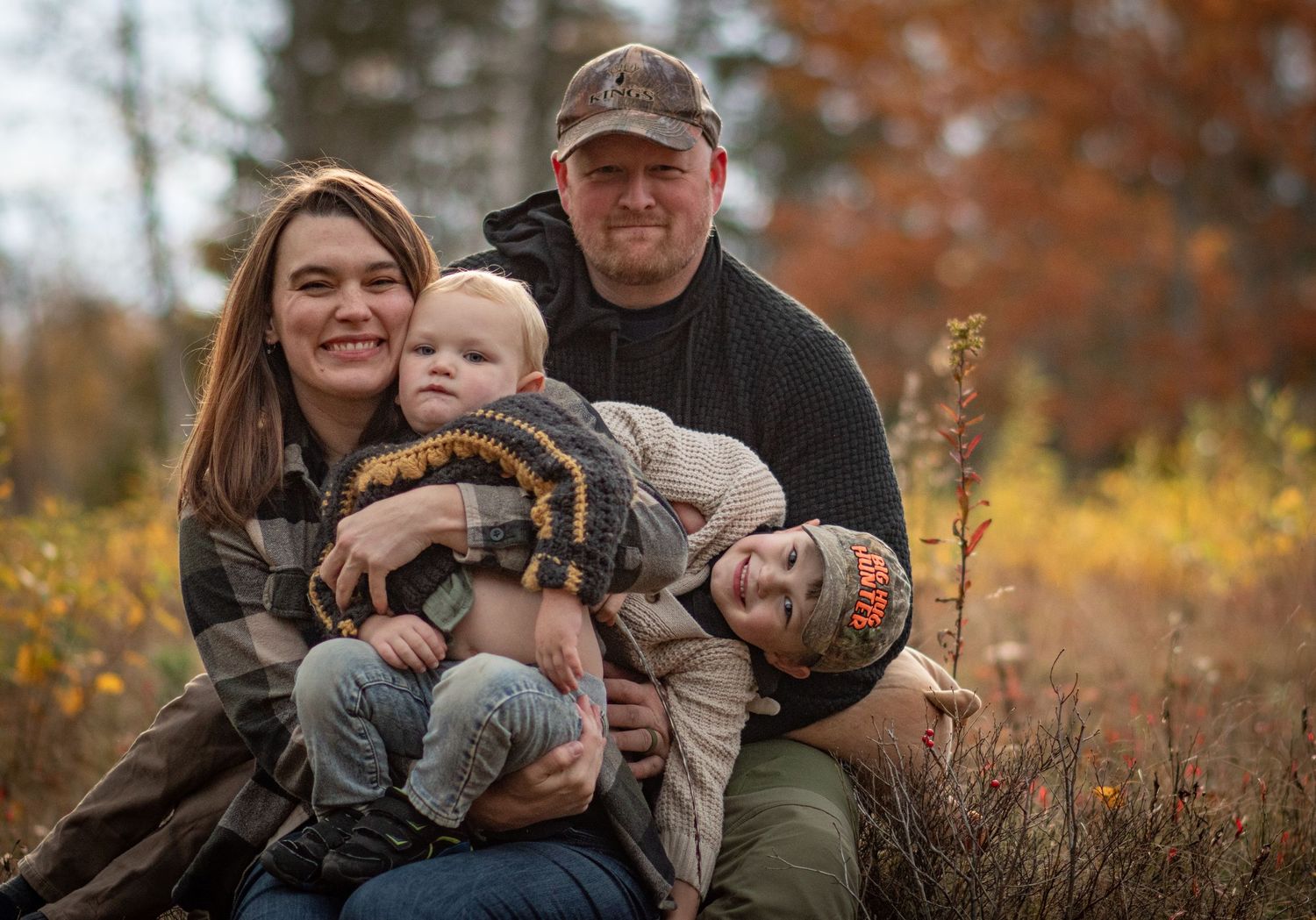 Smiling young family of four posing together in vibrant autumn forest setting. Photo by Xenophon Photography in Maine.