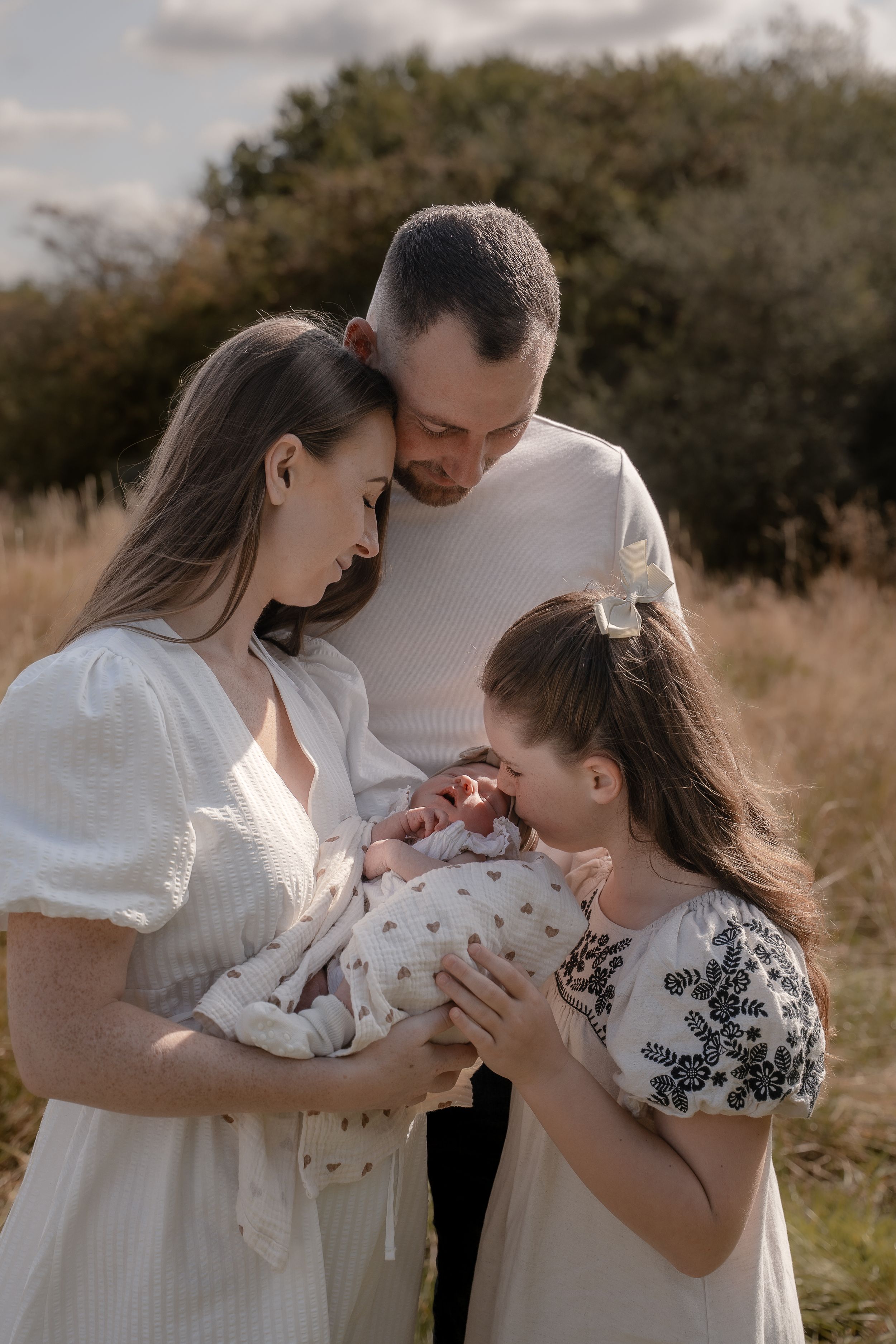 A family shares a tender moment outdoors while holding a newborn baby wrapped in polka dot swaddle in golden sunlight.