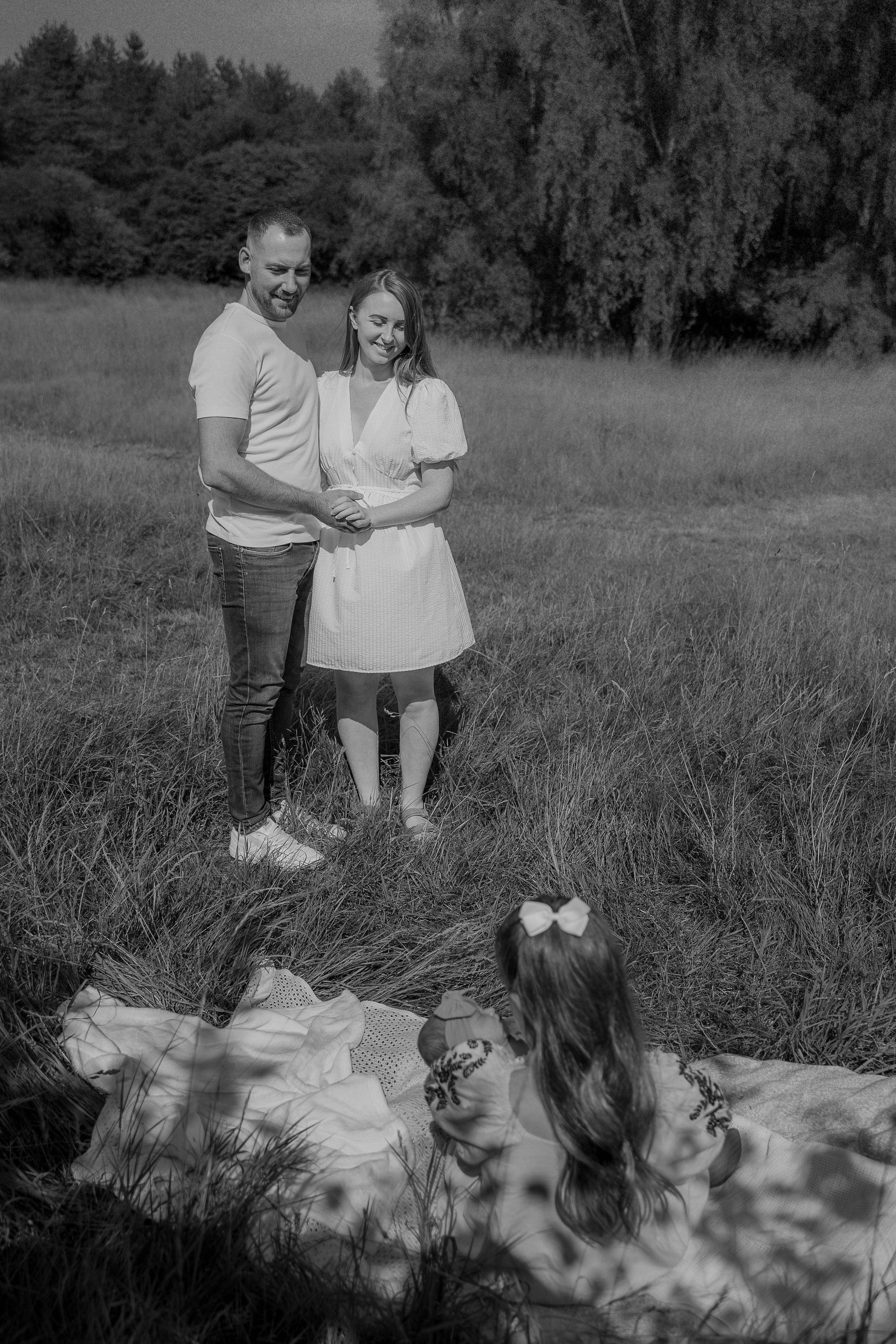 A black and white outdoor portrait of a couple standing in a grassy field with trees in the background.