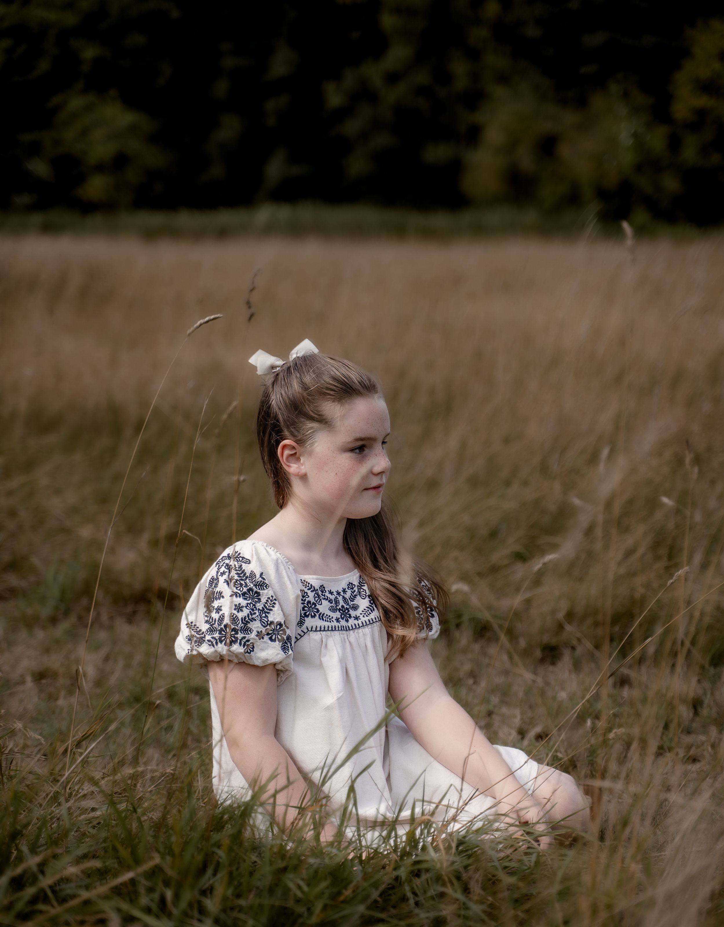 Young girl sitting in a meadow with long grass, wearing a cream dress with embroidered sleeves, captured in a Derbyshire family photography