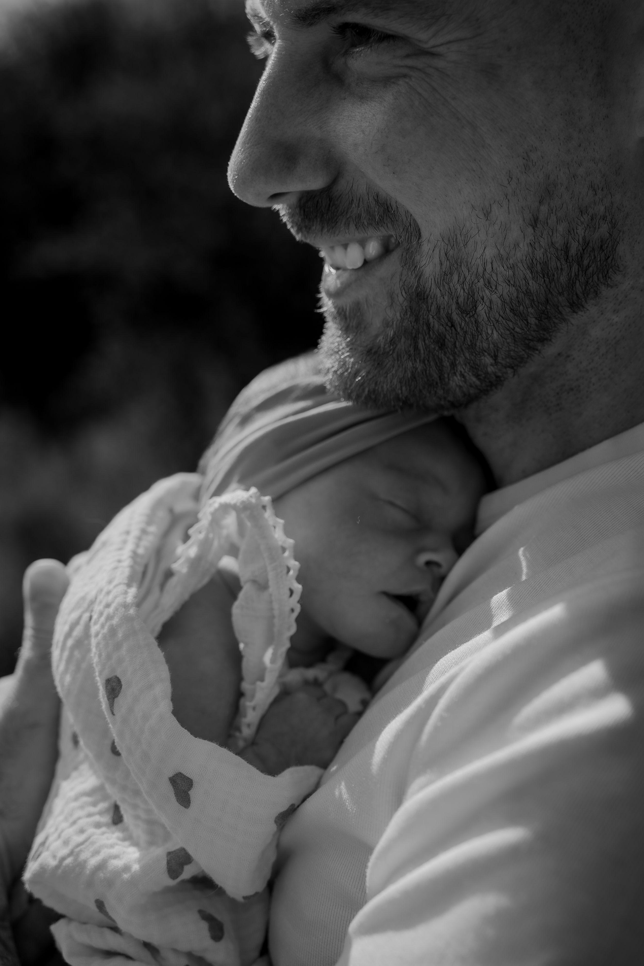 Father holding newborn baby close outdoors, smiling in a tender moment during a lifestyle photography session in Derbyshire.