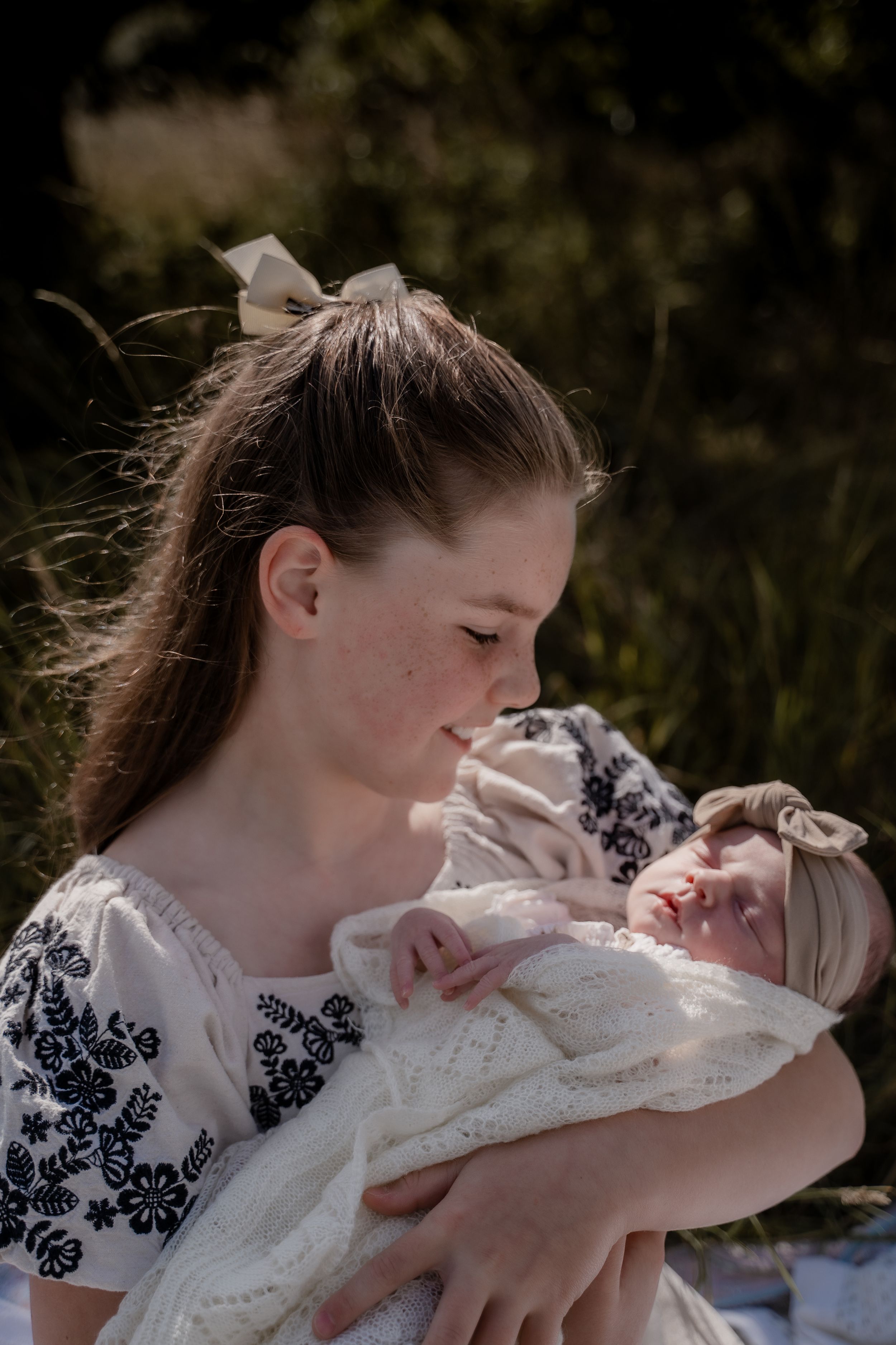 Older sister smiling down at her newborn sibling wrapped in a blanket during a warm outdoor lifestyle photography session in Derbyshire.