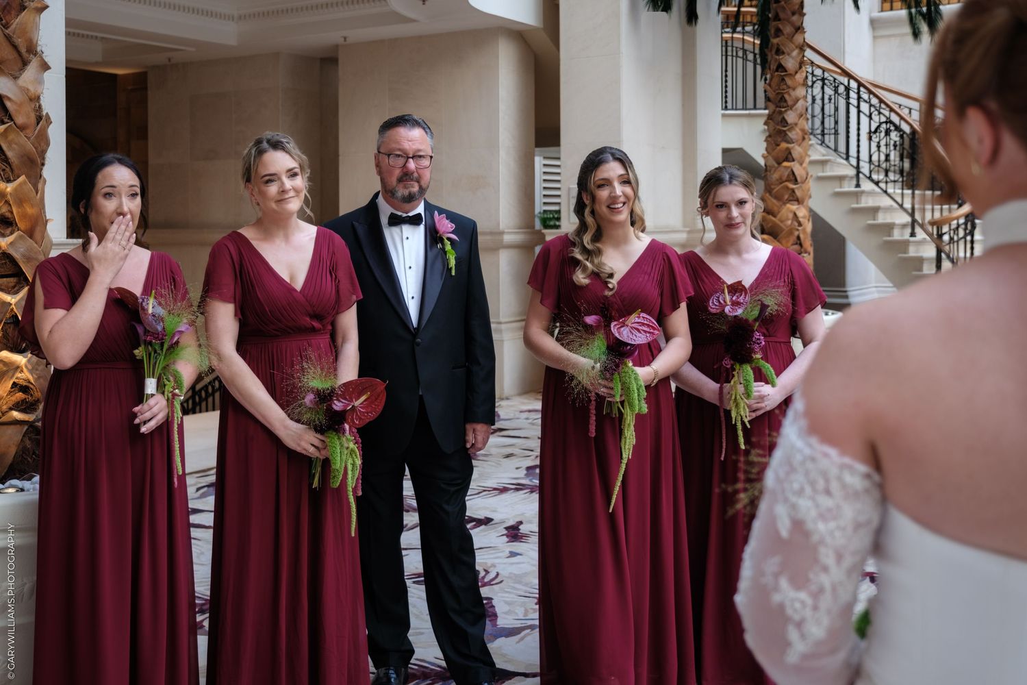 Wedding party members in burgundy dresses gather with others in formal attire at an elegant indoor venue.