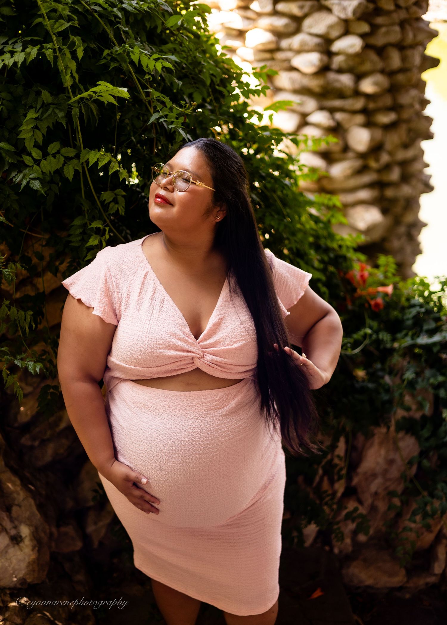 Pregnant woman in pink crop top and skirt poses outdoors among lush green foliage and stone wall.
