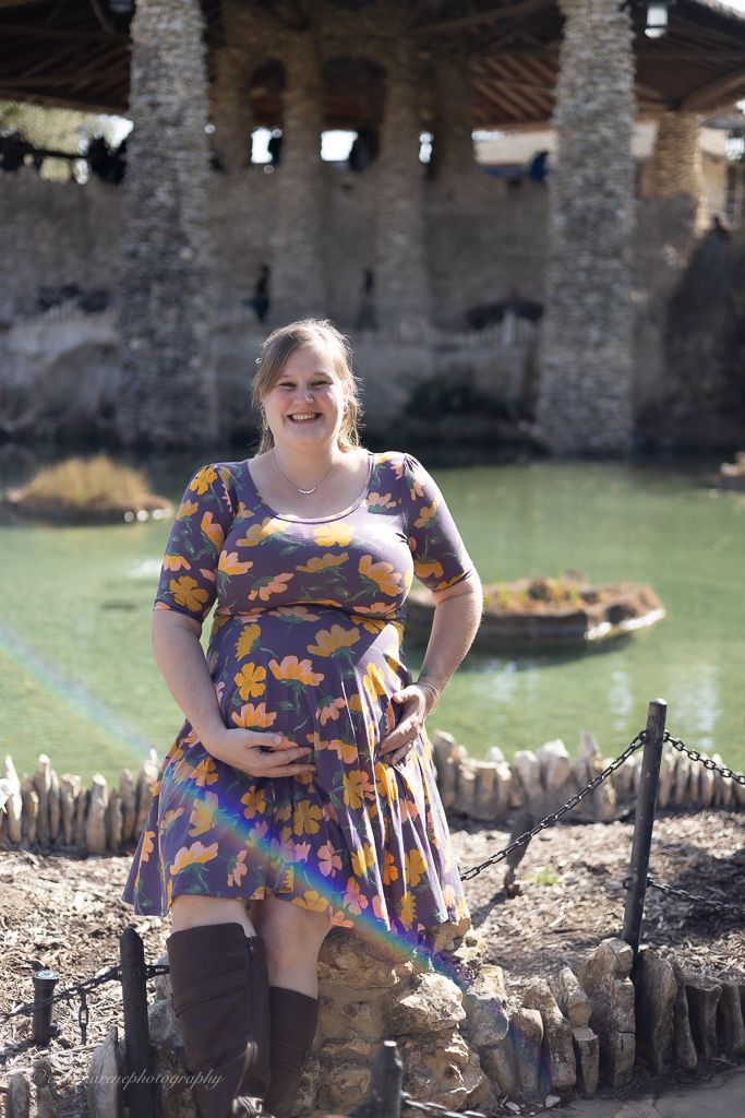 Smiling pregnant woman in floral purple dress holding belly, standing near stone fountain outdoors.