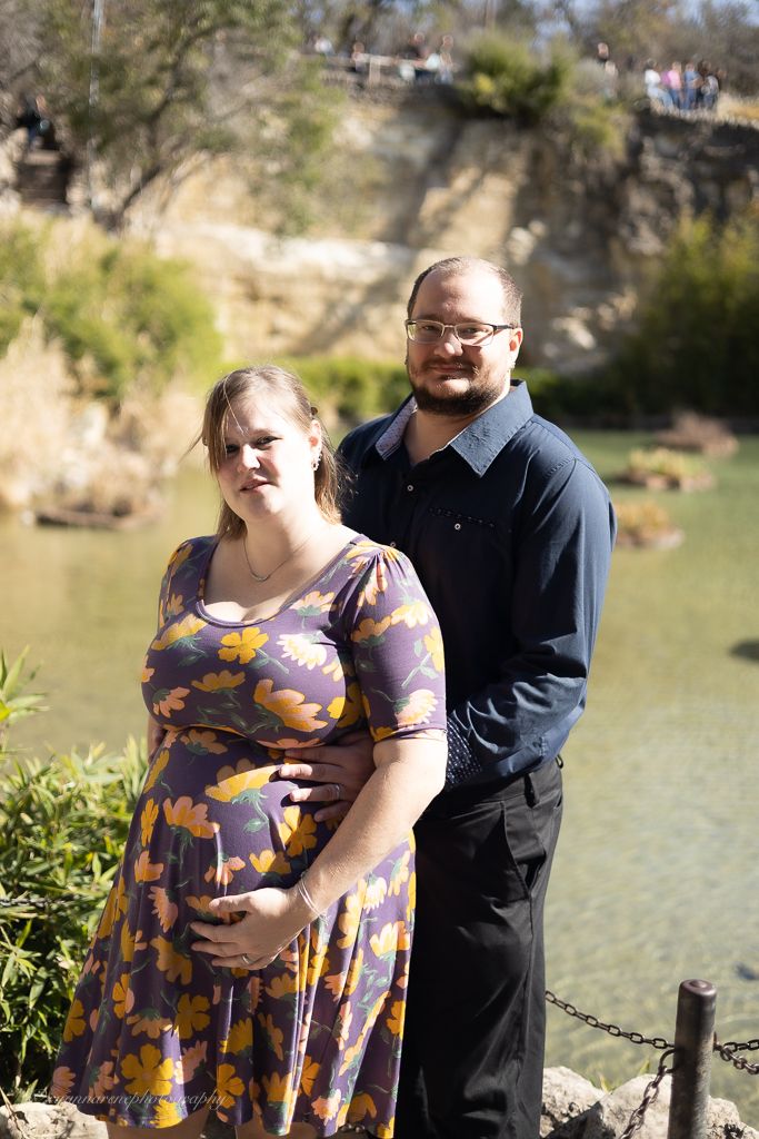 Pregnant woman in floral dress and man in navy shirt pose together outdoors near a scenic rocky waterway.